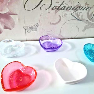 May include: Five heart-shaped decorative bowls in various colours: clear, purple, red, white, and turquoise. The bowls are arranged on a white surface, with a floral and butterfly-themed background.