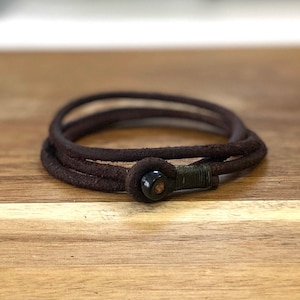 May include: A dark brown leather wrap bracelet with a black clasp. The bracelet is wrapped multiple times and has a rustic, worn appearance. The clasp is a dark, circular shape.