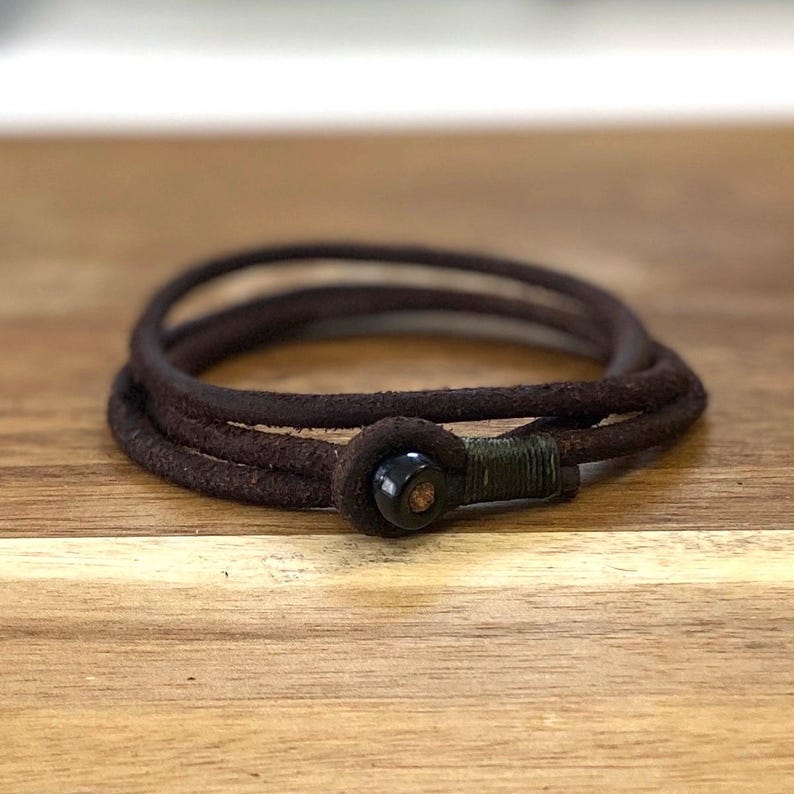 May include: A dark brown leather wrap bracelet with a black clasp. The bracelet is wrapped multiple times and has a rustic, worn appearance. The clasp is a dark, circular shape.