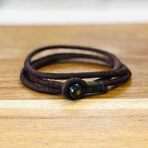 May include: A dark brown leather bracelet with a wrapped closure and a black bead. The bracelet is made of three strands of leather and is displayed on a wooden surface. The bracelet is a stylish accessory.