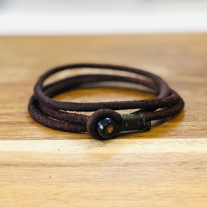 May include: A dark brown leather bracelet with a wrapped closure and a black bead. The bracelet is made of three strands of leather and is displayed on a wooden surface. The bracelet is a stylish accessory.