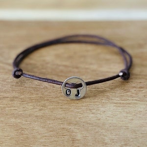 Puede incluir: Una pulsera de cuero marrón con un dije plateado. El dije tiene las iniciales "OJ". La pulsera está atada con un nudo. La pulsera mide aproximadamente 18 cm de largo.