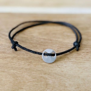 May include: A minimalist bracelet with a black cord and a small, circular silver-toned charm. The charm has a unique cutout design. The bracelet is displayed on a light brown wooden surface, showcasing its simple elegance.
