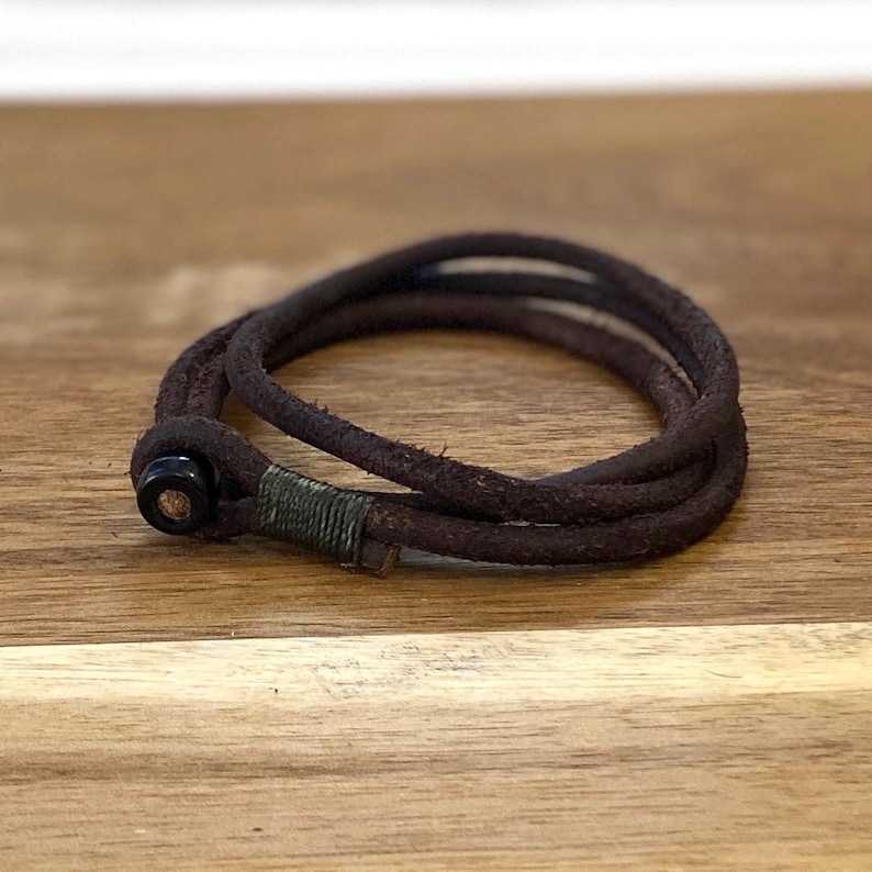 May include: A dark brown leather bracelet with a wrap-around design. The bracelet features a black clasp and a green wrapped accent. The bracelet is displayed on a wooden surface.