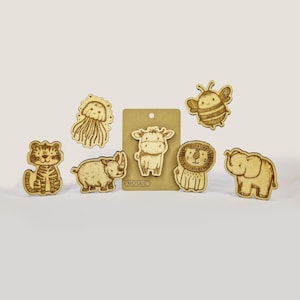 Könnte beinhalten: Satz mit sieben Tiermagneten aus Holz, darunter ein Tiger, ein Nashorn, eine Qualle, eine Kuh, eine Biene, ein Löwe und ein Elefant. Die Magnete sind alle braun und haben ein cartoonartiges Design. Die Magnete sind auf einem weißen Hintergrund mit einer braunen Karte in der Mitte angeordnet, auf der "MOSAIC" steht.