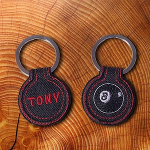 May include: Two black keychains with red stitching. One keychain has the word "TONY" embroidered on it. The other keychain has a white embroidered image of a black 8-ball.