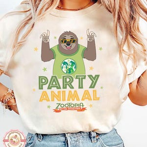 May include: Cream-coloured t-shirt with a cartoon sloth wearing sunglasses and a green shirt with a graphic. The shirt has the words "PARTY ANIMAL" in green and yellow, with "ZOOTOPIA" below. The design includes stars.