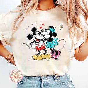 May include: Cream-colored t-shirt featuring a cartoon illustration of Mickey and Minnie Mouse embracing. Mickey wears red shorts and yellow shoes, while Minnie dons a blue polka-dot dress and pink shoes. Hearts and a flower accent the design, evoking a romantic theme.
