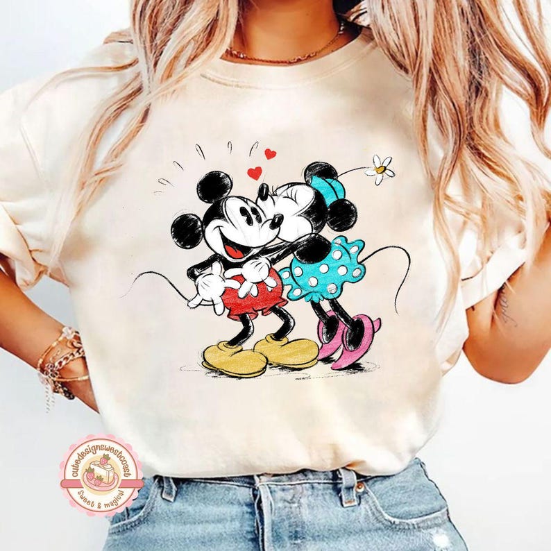 May include: Cream-colored t-shirt featuring a cartoon illustration of Mickey and Minnie Mouse embracing. Mickey wears red shorts and yellow shoes, while Minnie dons a blue polka-dot dress and pink shoes. Hearts and a flower accent the design, evoking a romantic theme.