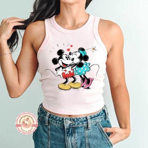 May include: A light pink tank top featuring a graphic of Mickey and Minnie Mouse embracing, with hearts and a flower. The cartoon characters are in their classic outfits. The tank top has a rounded neckline and armholes.