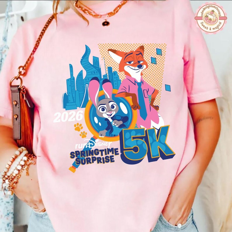 May include: Pale pink t-shirt with a Zootopia design. It features Judy Hopps and Nick Wilde, the text "2026", "5K", and "Springtime Surprise". The top has a crew neck and short sleeves.