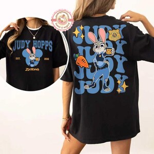 May include: Black t-shirt featuring Judy Hopps from Zootopia. The front design includes "JUDY HOPPS" in blue, a cartoon image of Judy, and "EST. 2016". The back displays "JUDY" repeated with Judy Hopps, a carrot, and police badge.