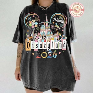 May include: Black t-shirt featuring a colorful Disneyland design with the text "Disneyland 2026". The design includes Mickey Mouse ears, a castle, fireworks, and various Disney characters. The shirt is a relaxed fit.