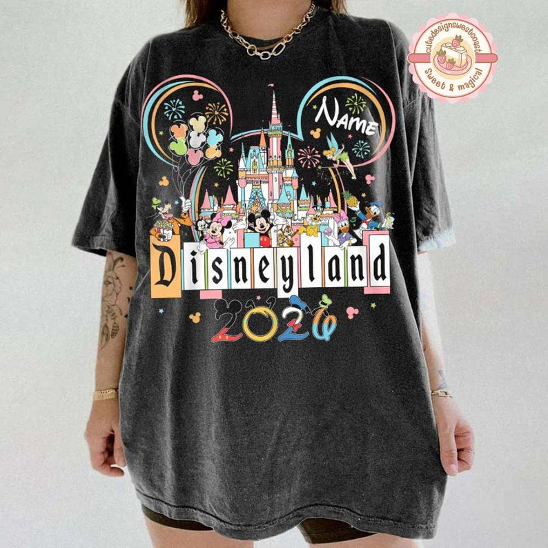 May include: Black t-shirt featuring a colorful Disneyland design with the text "Disneyland 2026". The design includes Mickey Mouse ears, a castle, fireworks, and various Disney characters. The shirt is a relaxed fit.