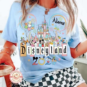 May include: Light blue t-shirt featuring a Disneyland graphic. The design includes a castle, Disney characters, and the text "Disneyland 2020". Colorful balloons and fireworks are also present in the design.