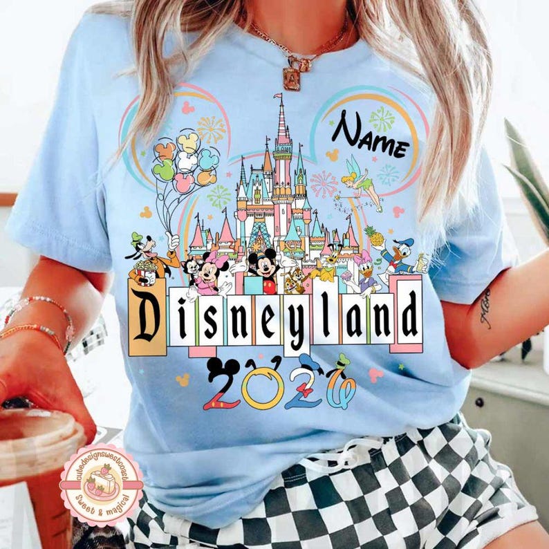 May include: Light blue t-shirt featuring a Disneyland graphic. The design includes a castle, Disney characters, and the text "Disneyland 2020". Colorful balloons and fireworks are also present in the design.