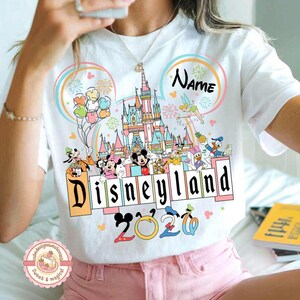 May include: White t-shirt featuring a colorful Disneyland graphic with Mickey Mouse, Minnie Mouse, and other Disney characters. The design includes a castle, fireworks, and the text "Disneyland 2020". A name can be added above the castle.