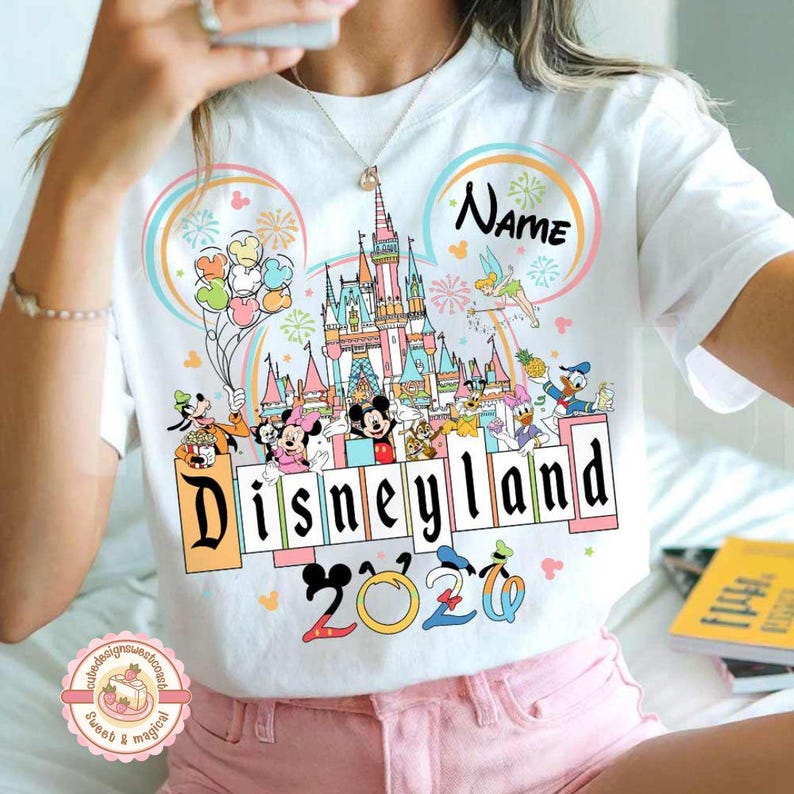 May include: White t-shirt featuring a colorful Disneyland graphic with Mickey Mouse, Minnie Mouse, and other Disney characters. The design includes a castle, fireworks, and the text "Disneyland 2020". A name can be added above the castle.