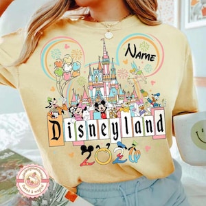 May include: Yellow Disneyland 2020 sweatshirt featuring a colorful castle graphic, Disney characters, and the word "Disneyland" in a block letter design. The sweatshirt has a round neckline and long sleeves.