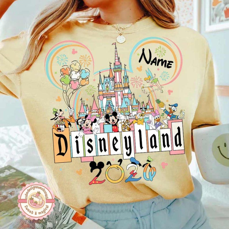 May include: Yellow Disneyland 2020 sweatshirt featuring a colorful castle graphic, Disney characters, and the word "Disneyland" in a block letter design. The sweatshirt has a round neckline and long sleeves.