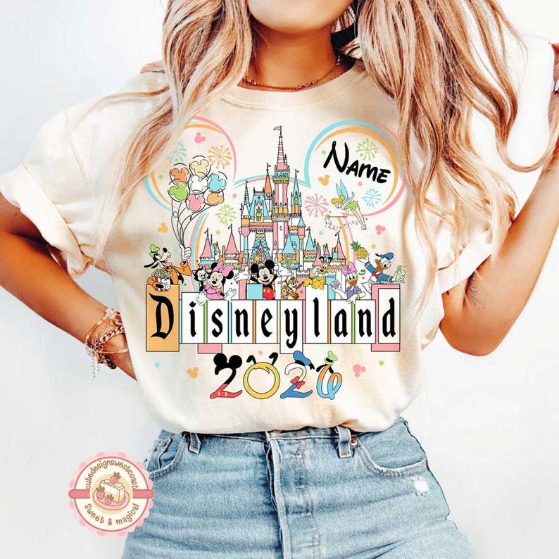 May include: Cream-colored t-shirt featuring a colorful Disneyland design. The design includes a castle, Disney characters, balloons, and the text "Disneyland" and "2020". A space for a name is also included.