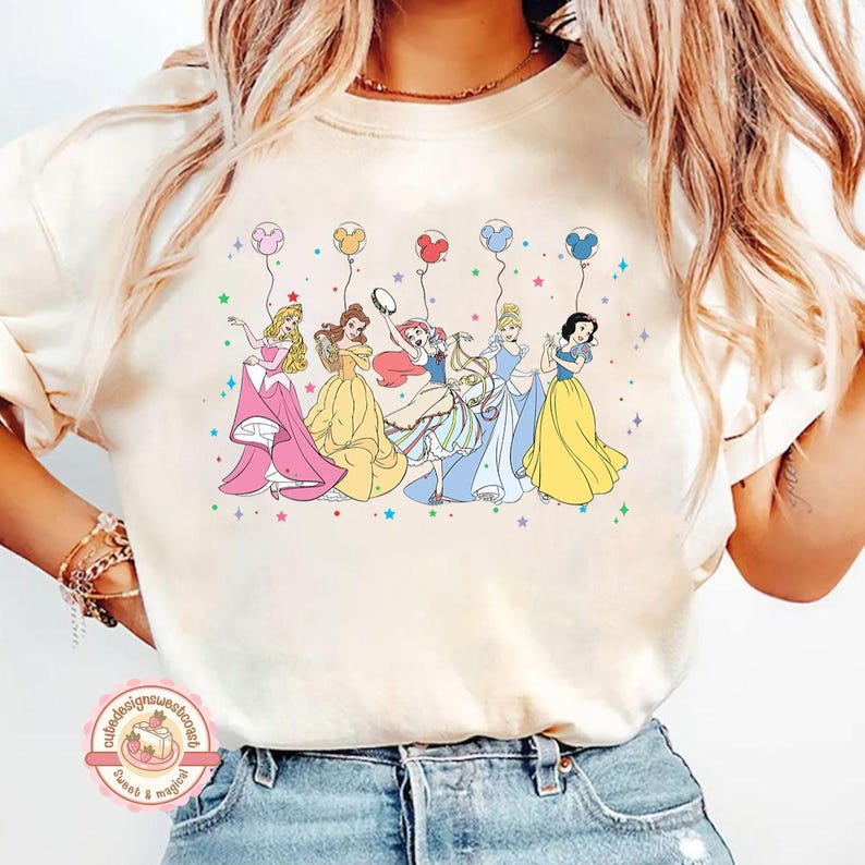 May include: A white t-shirt with a graphic of six Disney princesses holding balloons. The princesses are Snow White, Cinderella, Belle, Ariel, Aurora, and Rapunzel. The balloons are in various colors, including red, blue, green, yellow, and purple.