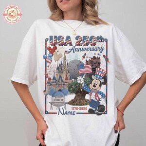 May include: White t-shirt featuring a colorful graphic celebrating the USA's 250th Anniversary. The design includes Mickey Mouse in patriotic attire, iconic landmarks, fireworks, and the text "USA 250th Anniversary 1776-2026".