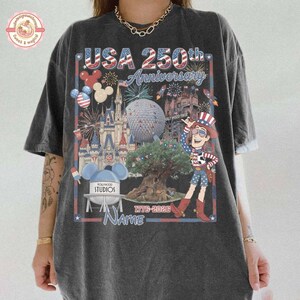 May include: Dark gray t-shirt with a colorful graphic celebrating the USA's 250th Anniversary. The design includes fireworks, a cartoon character, and various Disney park landmarks. The text "USA 250th Anniversary" is prominently displayed.