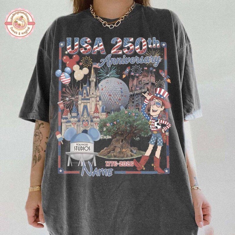 May include: Dark gray t-shirt with a colorful graphic celebrating the USA's 250th Anniversary. The design includes fireworks, a cartoon character, and various Disney park landmarks. The text "USA 250th Anniversary" is prominently displayed.