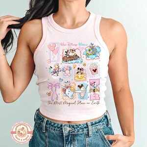 May include: A light pink tank top with a colorful Disney World graphic. The graphic includes images of Mickey and Minnie Mouse, Dumbo, and other Disney characters. The text "The Most Magical Place on Earth" is also visible.