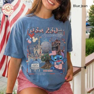 May include: A light blue t-shirt with a graphic celebrating the USA's 250th Anniversary. The design includes the text "USA 250th Anniversary", fireworks, and a cartoon character holding an American flag. The shirt also features the text "1776-2026".
