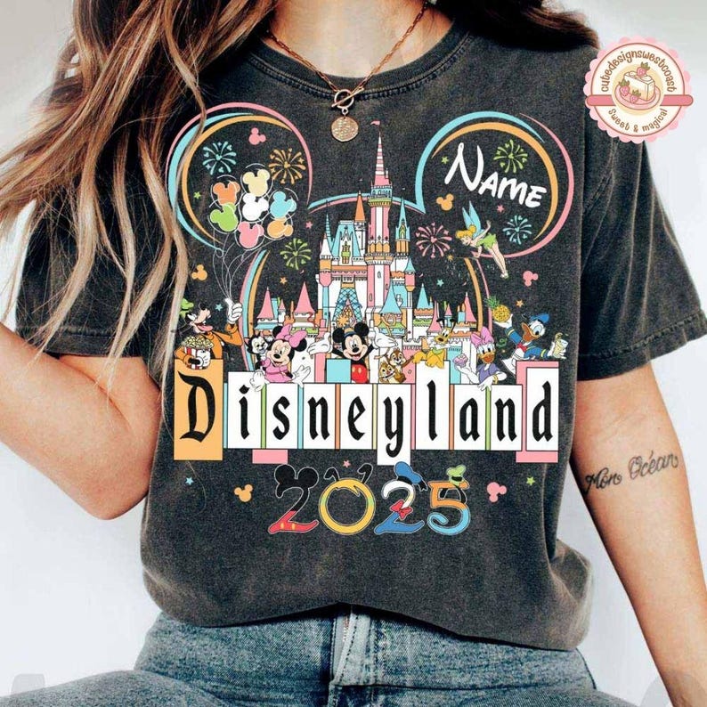 May include: A black t-shirt with a vintage-style graphic of Mickey Mouse's ears, Cinderella's castle, and fireworks. The text "Disneyland 2025" is printed below the graphic. The text "Name" is printed in the upper right corner of the graphic.