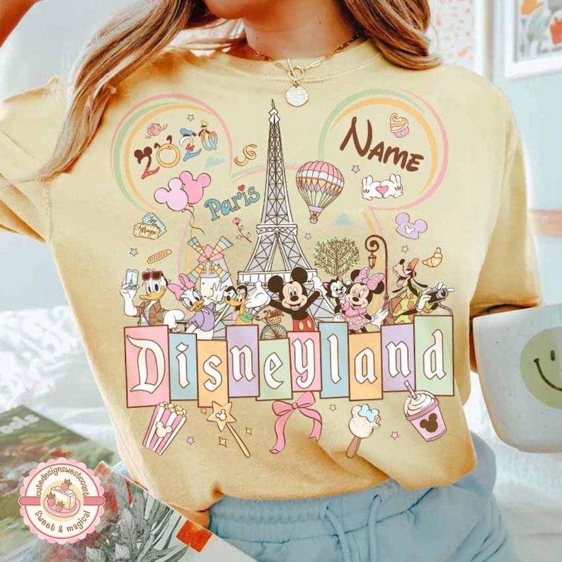 May include: Yellow t-shirt with a Disneyland Paris design. The graphic includes the Eiffel Tower, Mickey and Minnie Mouse, Donald and Daisy Duck, and other Disney characters. The word "Disneyland" is displayed in colorful letters.