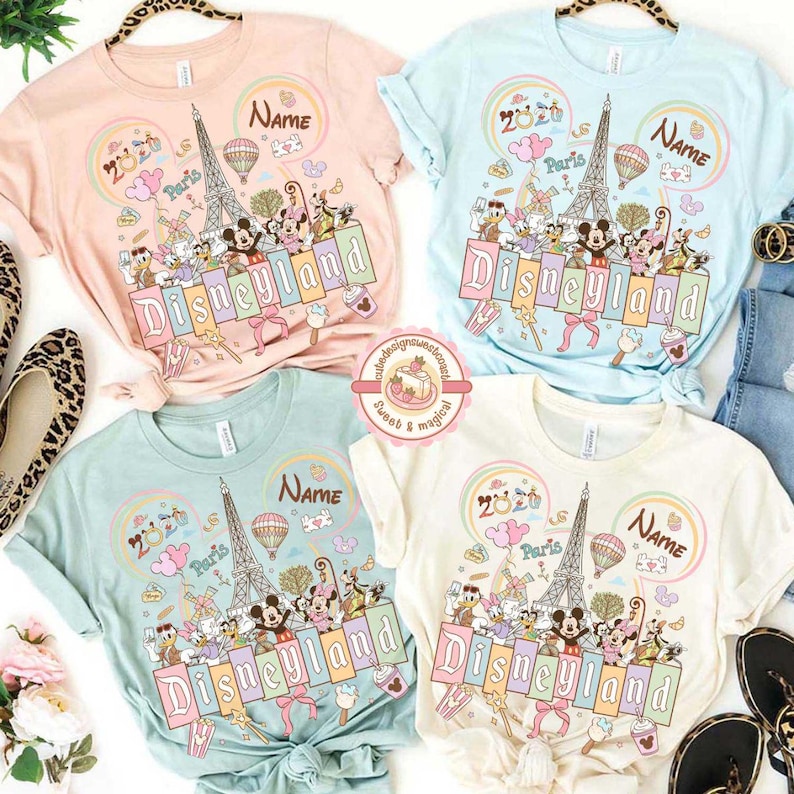 May include: Four pastel-colored t-shirts with a Disneyland theme. Each shirt features a graphic with the Eiffel Tower, Mickey Mouse, and other Disney characters. The word "Disneyland" is prominently displayed.