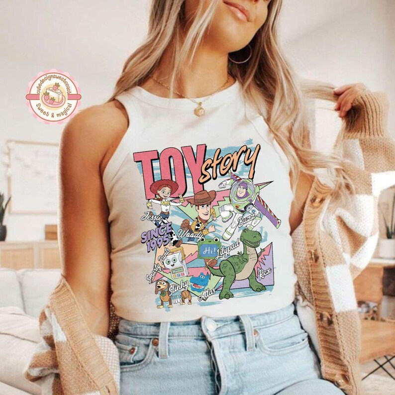 Pu&ograve; includere: Una canotta senza maniche color crema con una stampa grafica colorata che include il testo TOY story, Jessie, Woody, Buzz, Slinky, Rex e altri personaggi. Il design presenta varie forme geometriche e il testo SINCE 1995 in stile retro.
