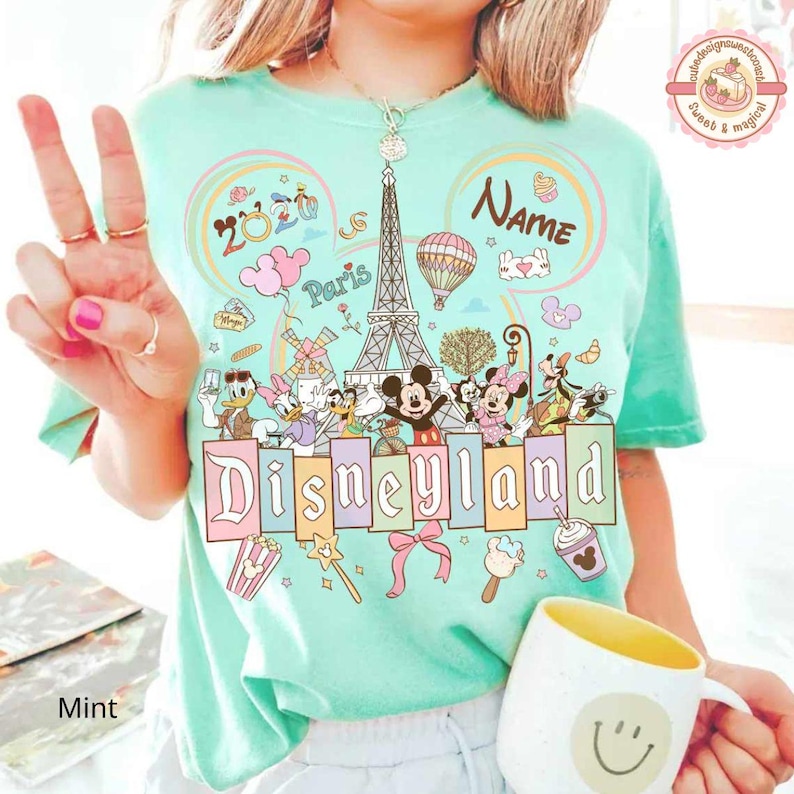 May include: Mint green t-shirt featuring the word "Disneyland" in pastel colors, with illustrations of Disney characters and the Eiffel Tower. The shirt includes the text "Paris" and "2023". A yellow and white mug with a smiley face is in the foreground.