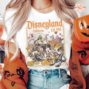K&ouml;nnte beinhalten: Wei&szlig;es T-Shirt mit einem Disneyland California-Design, mit dem Text "Disneyland California Est. 1955". Die Grafik zeigt Cartoon-Figuren als Skelette. Das Shirt hat ein Vintage-Halloween-Thema.