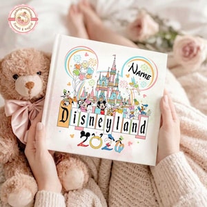 May include: A white Disneyland-themed photo album with a colorful illustration of the Disneyland castle, characters, and fireworks. The album has the word "Disneyland" and the year "2020" printed on it. A teddy bear is next to the album.