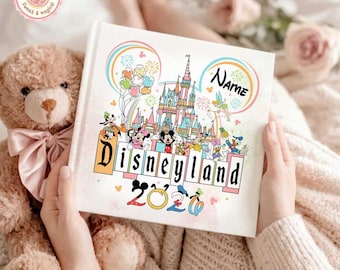 May include: A white Disneyland-themed photo album with a colorful illustration of the Disneyland castle, characters, and fireworks. The album has the word "Disneyland" and the year "2020" printed on it. A teddy bear is next to the album.