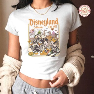 K&ouml;nnte beinhalten: Wei&szlig;es T-Shirt mit einem Disneyland Halloween-Design. Die Grafik zeigt Micky Maus, Minnie Maus, Donald Duck und Goofy in Skelett-Kost&uuml;men, mit dem Text "Disneyland California Est. 1955". Das Design ist vor dem Hintergrund von Parkbildern platziert.