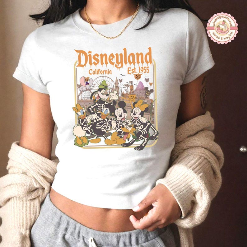 K&ouml;nnte beinhalten: Wei&szlig;es T-Shirt mit einem Disneyland Halloween-Design. Die Grafik zeigt Micky Maus, Minnie Maus, Donald Duck und Goofy in Skelett-Kost&uuml;men, mit dem Text "Disneyland California Est. 1955". Das Design ist vor dem Hintergrund von Parkbildern platziert.