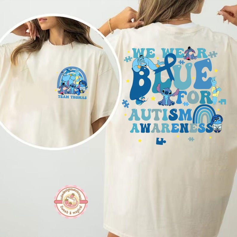 May include: Cream-coloured t-shirt with a graphic on the front and back. The front features a blue cartoon character design with the text "TEAM THOMAS". The back displays the text "WE WEAR BLUE FOR AUTISM AWARENESS" with cartoon characters.