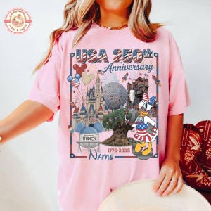 May include: Pink t-shirt with a graphic celebrating the USA's 250th anniversary. The design includes Disney park icons, fireworks, and a patriotic Daisy Duck. The text reads "USA 250th Anniversary" and "1776-2026".