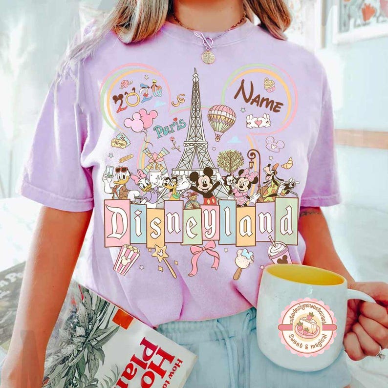 May include: A light purple t-shirt with a Disneyland graphic featuring Mickey and Minnie Mouse, Donald Duck, and other cartoon characters. The shirt also has the Eiffel Tower and the word "Paris".