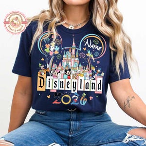 May include: Navy blue t-shirt featuring a colorful Disneyland graphic. The design includes the Disneyland castle, Mickey Mouse, and other characters. The text "Disneyland 2020" is also displayed.