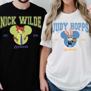 May include: Black and white t-shirts featuring characters from the animated film Zootopia. One shirt displays "Nick Wilde" with a fox illustration, the other "Judy Hopps" with a bunny illustration. Both shirts have "Zootopia" text and the year "2016".