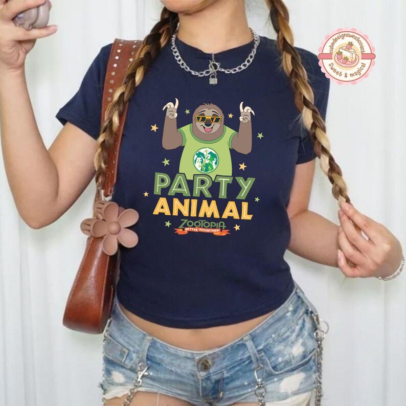 May include: A navy blue t-shirt with a sloth wearing sunglasses and a green shirt with a globe design. The text "PARTY ANIMAL" and "ZOOTOPIA" are printed below the sloth. The shirt is worn with denim shorts.