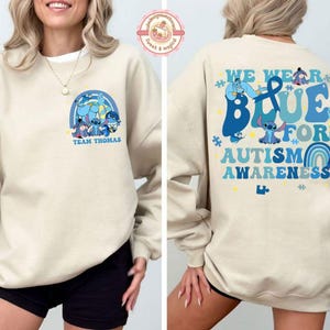May include: A cream-coloured sweatshirt with a blue graphic on the front featuring cartoon characters and the text "Team Thomas". The back displays the message "We Wear Blue for Autism Awareness" in blue, with more cartoon characters.