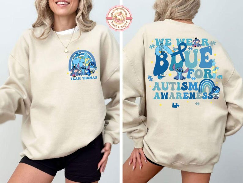 May include: A cream-coloured sweatshirt with a blue graphic on the front featuring cartoon characters and the text "Team Thomas". The back displays the message "We Wear Blue for Autism Awareness" in blue, with more cartoon characters.