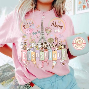 May include: Pink Disneyland t-shirt with a colorful graphic featuring Mickey and Minnie Mouse, Donald and Daisy Duck, and other Disney characters. The design includes the Eiffel Tower, a hot air balloon, and the word "Disneyland". A light blue mug with a dessert design is also visible.
